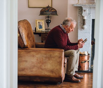 A senior adult man sitting alone in his living room looking sad as he holds a photo in his hands